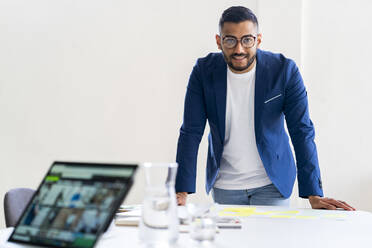 Smiling businessman with eyeglasses leaning on desk at office - GIOF12311