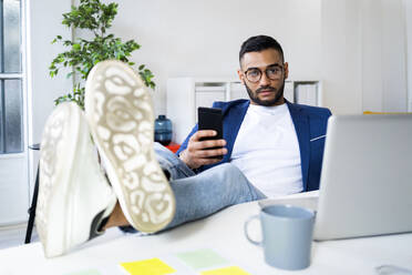Businessman with smart phone relaxing with feet up on desk at office - GIOF12315