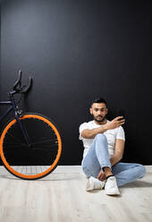 Junger Mann mit Smartphone sitzt in der Nähe des Fahrrads an der Wand im Studio - GIOF12325