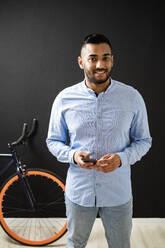 Smiling man with smart phone standing by bicycle on wall in background - GIOF12326