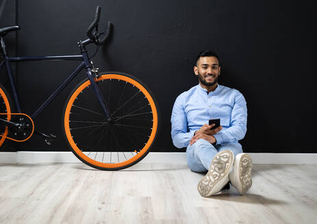 Handsome man with smart phone sitting near bicycle by wall at studio - GIOF12327