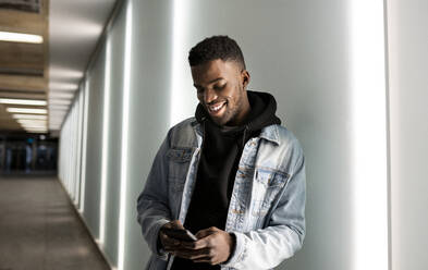 Smiling young man using smart phone while standing on wall - JCCMF01815