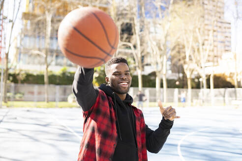 Cheerful man showing shaka sign while playing basketball on sunny day - JCCMF01817
