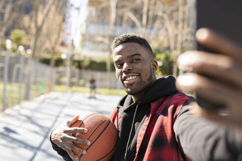 Junger Mann hält Basketball, während er ein Selfie mit seinem Handy an einem sonnigen Tag macht - JCCMF01819