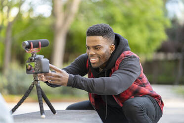 Smiling male vlogger adjusting camera on tripod at public park - JCCMF01824