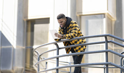 Junger Mann, der ein digitales Tablet benutzt und sich auf ein Geländer stützt - JCCMF01849