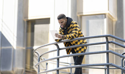 Young man using digital tablet while leaning on railing - JCCMF01849