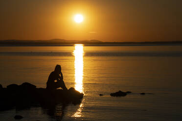 Silhouette eines Teenagers, der auf einem Felsen sitzt und den Sonnenuntergang über dem Trasimenischen See, Umbrien, Italien, betrachtet - LOMF01283
