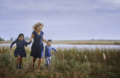 Mother holding hands while running with daughter and son on grass - AZF00272