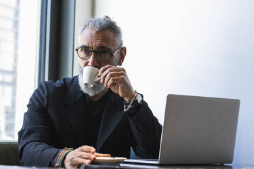 Geschäftsmann trinkt Kaffee, während er in einem Café vor einem Laptop sitzt - PNAF01331