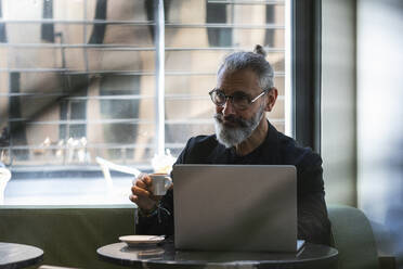 Smiling hipster male entrepreneur contemplating while having coffee cup in shop - PNAF01332