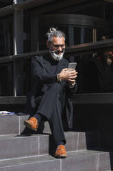 Lächelnder männlicher Berufstätiger, der ein Mobiltelefon benutzt, während er auf den Stufen eines Gebäudes sitzt - PNAF01343
