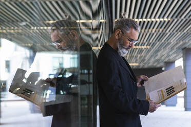 Hipster businessman reading business plan while standing in front of glass wall - PNAF01348