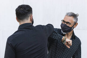 Geschäftsmann mit Schutzmaske, der mit einem männlichen Kollegen vor einer Wand den Ellbogen anstößt - PNAF01386
