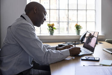 Männlicher Berufstätiger, der auf einem Laptop eine Haftnotiz schreibt, während er am Schreibtisch im Büro sitzt - WPEF04237