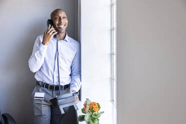 Lächelnder männlicher Unternehmer, der am Festnetztelefon spricht und sich im Büro an die Wand lehnt - WPEF04241