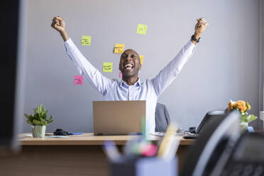 Cheerful businessman with arms raised while sitting with laptop at office - WPEF04257