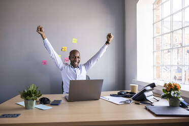 Businessman with hands raised looking at laptop on desk at office - WPEF04258