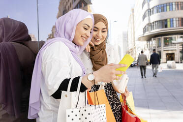 Arabische Freundinnen benutzen ein Smartphone, während sie sich an eine Glaswand in der Stadt lehnen - JCCMF01866