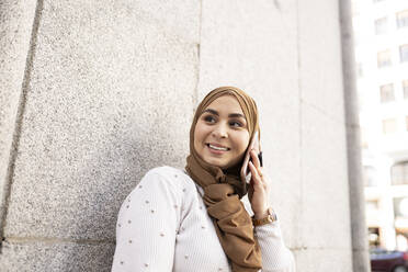 Junge Frau im Hidschab schaut weg, während sie mit einem Smartphone an der Wand telefoniert - JCCMF01888