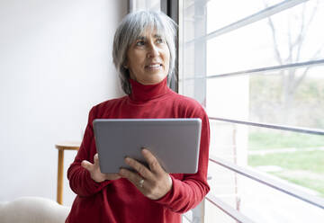 Lächelnde reife Frau, die wegschaut, während sie ein digitales Tablet zu Hause hält - JCCMF01894