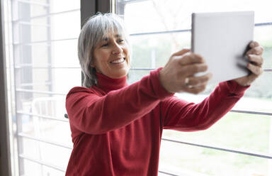 Lächelnde Frau nimmt Selfie durch digitale Tablet durch Fenster zu Hause - JCCMF01895