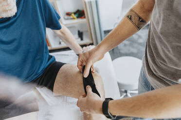 Männlicher Physiotherapeut klebt elastisches therapeutisches Band auf das Knie eines Patienten - MFF07707