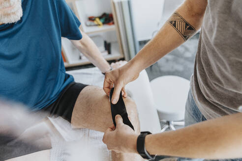 Männlicher Physiotherapeut klebt elastisches therapeutisches Band auf das Knie eines Patienten - MFF07707