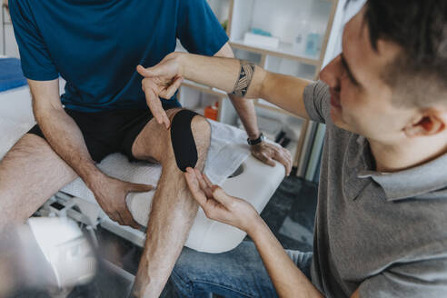 Männlicher Physiotherapeut klebt Kinesio-Tape auf das Knie eines Patienten am Massagetisch in der Praxis - MFF07710