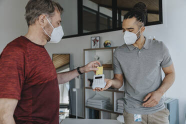 Physiotherapist in protective face mask holding credit card reader while patient paying in medical practice - MFF07780