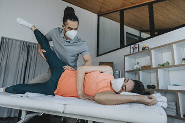 Male medical professional wearing face mask stretching leg of patient during physiotherapy in medical practice - MFF07809