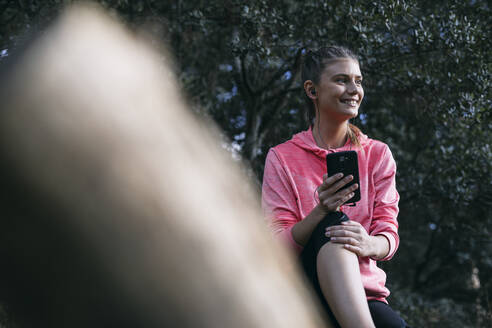 Lächelnde Frau, die wegschaut, während sie über ihr Smartphone im Wald Musik hört - ABZF03531