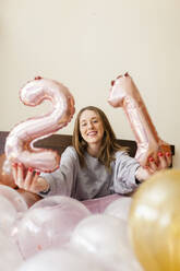 Lächelnde Frau hält Nummer 21 Heliumballons, während sie auf dem Bett zu Hause sitzt - MRRF01014