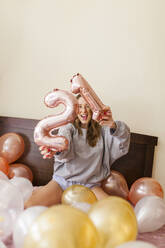 Cheerful woman with number 21 helium balloons sitting on bed at home - MRRF01015