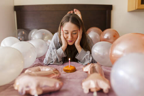 Frau mit Kopf in den Händen, die auf eine Kerze auf einem Krapfen schaut, während sie inmitten von Luftballons liegt - MRRF01017