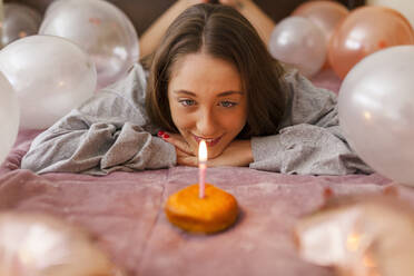 Junge Frau betrachtet eine brennende Kerze auf einem Krapfen, während sie zu Hause auf dem Bett liegt - MRRF01018