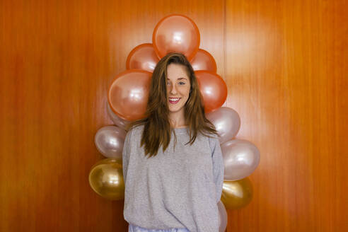 Smiling woman standing by wall with balloons decoration at home - MRRF01020