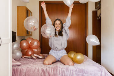 Fröhliche Frau spielt mit Luftballons, während sie zu Hause auf dem Bett kniet - MRRF01025