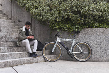 Junger Mann mit Fahrrad, der sein Smartphone benutzt, während er auf einer Treppe sitzt - JAQF00463