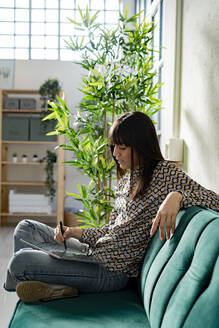 Weibliche Grafikdesignerin benutzt ein Tablet, während sie auf dem Sofa im Büro sitzt - GIOF12365