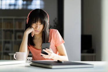 Frau mit Kopfhörern hält Smartphone, während sie zu Hause auf dem Tisch sitzt - GIOF12369