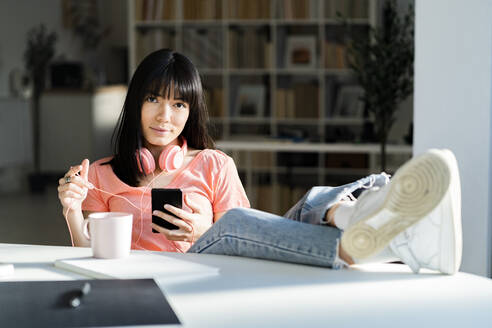Junge Frau mit hochgelegten Füßen auf dem Tisch, die ihr Smartphone zu Hause hält - GIOF12371