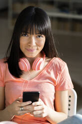 Smiling young woman with headphones holding smart phone at home - GIOF12372