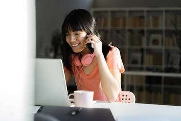 Smiling businesswoman talking on smart phone while sitting with laptop at home office - GIOF12378