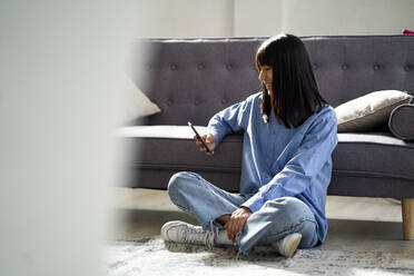 Smiling woman using smart phone while sitting on floor by sofa at home - GIOF12387