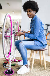 Afro hairstyle woman sitting on stool repairing bicycle at home - GIOF12398