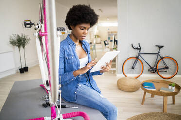 Afro-Frau sieht sich eine Anleitung auf einem digitalen Tablet an, während sie zu Hause ein Fahrrad repariert - GIOF12404