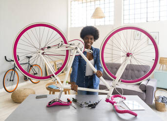 Smiling young woman holding bicycle on table while repairing at home - GIOF12407