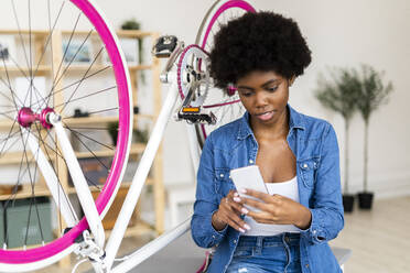 Young woman using mobile phone while sitting by bicycle on table at home - GIOF12413