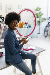 Smiling woman using mobile phone while sitting by bicycle on table at home - GIOF12415